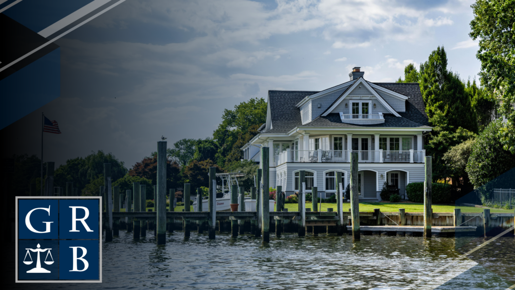 A white, multi-story waterfront house with a wraparound porch, facing a calm marina with pilings, under a partly cloudy sky, with a dark blue logo featuring GR and scales in the lower-left corner.